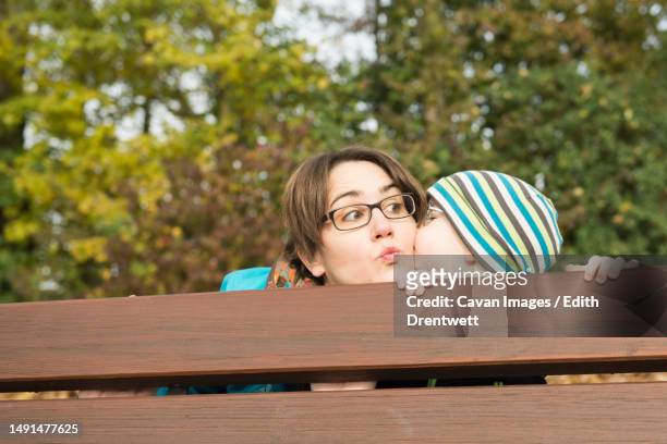 son giving his mother kiss on her cheek behind the backrest of park bench in autumn scenery - edith falls stock pictures, royalty-free photos & images