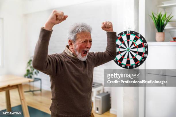 a senior man is playing darts at his home apartment and celebrating the victory. - man darts stock pictures, royalty-free photos & images