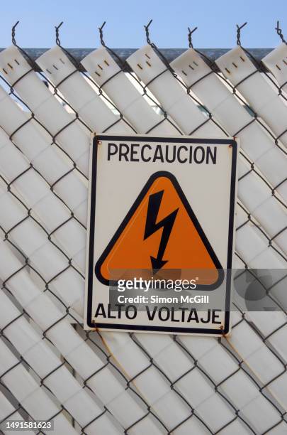 spanish-language 'precaución: alto voltaje [caution: high voltage]' warning sign on a chain-link fence - chain link fence on white stock-fotos und bilder