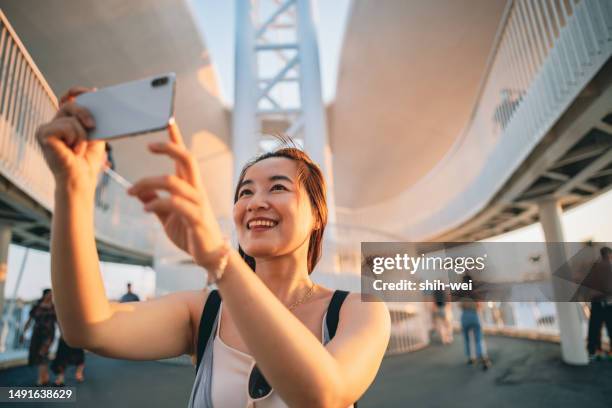 an asian woman fully embraces and enjoys her journey, using her smartphone to take selfies and record her memories at the tourist destination. - foreign born stock pictures, royalty-free photos & images