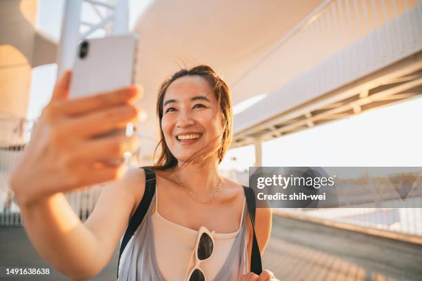 an asian woman fully embraces and enjoys her journey, using her smartphone to take selfies and record her memories at the tourist destination. - foreign born stock pictures, royalty-free photos & images