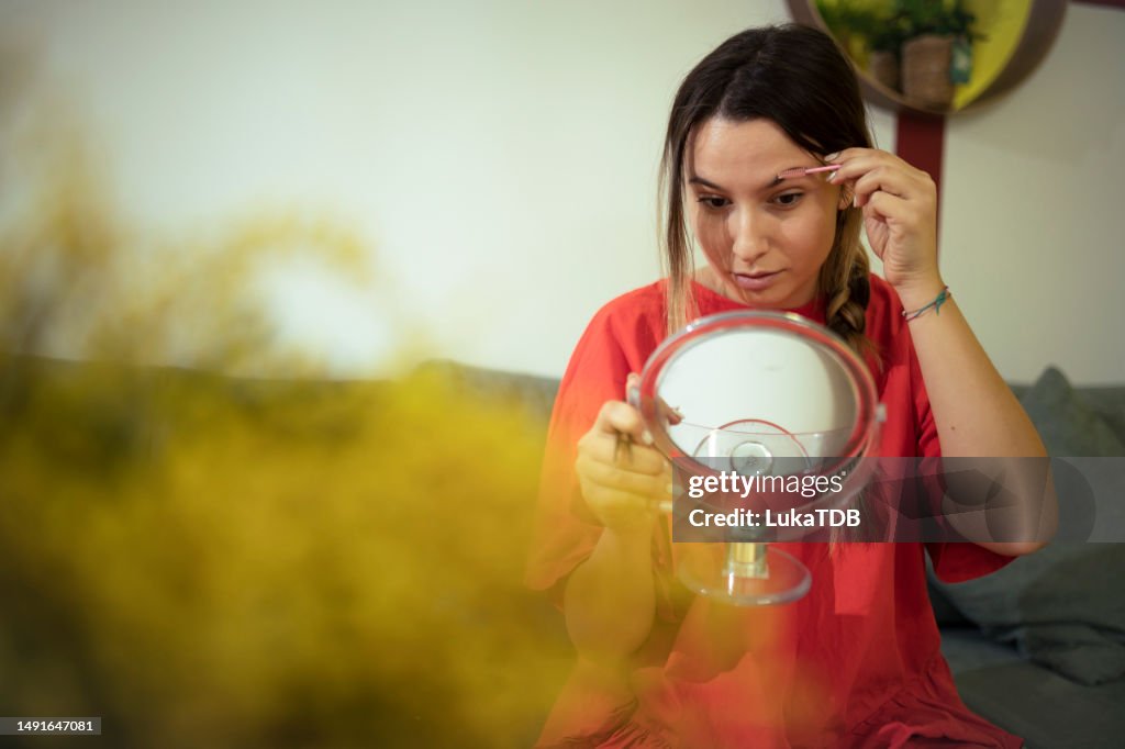 Beautiful woman tweezing eyebrows, looking in mirror