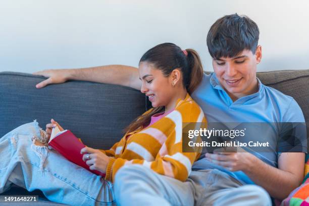 couple reading a book and using mobile phone while relaxing on the sofa at home. - casa calvet stock pictures, royalty-free photos & images