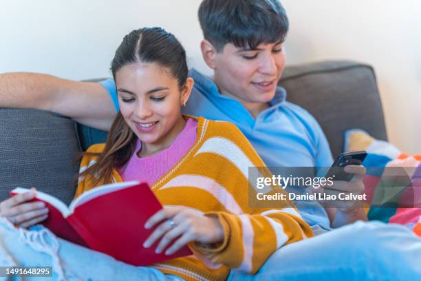 young couple relaxing on the sofa with a book and mobile phone. - casa calvet stock pictures, royalty-free photos & images