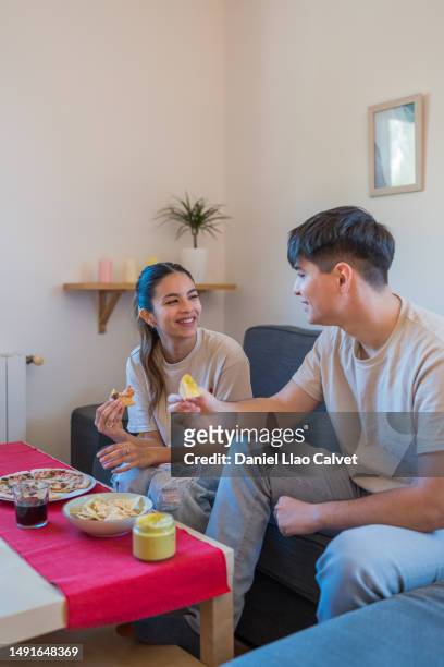 young couple eating nachos and pizza on the sofa at home. - casa calvet stock pictures, royalty-free photos & images
