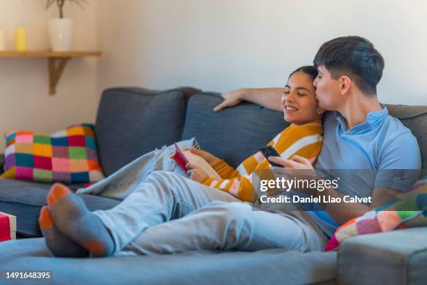 young man kissing his girlfriend while they relax on the couch in the living room at home. - casa calvet stock pictures, royalty-free photos & images
