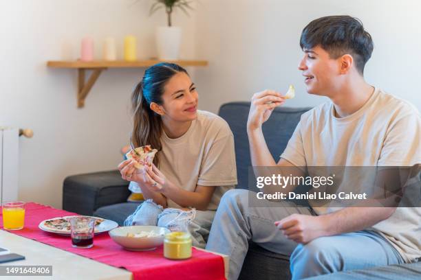 happy young couple enjoying eating nachos and pizza sitting on a sofa at home. - casa calvet stock pictures, royalty-free photos & images
