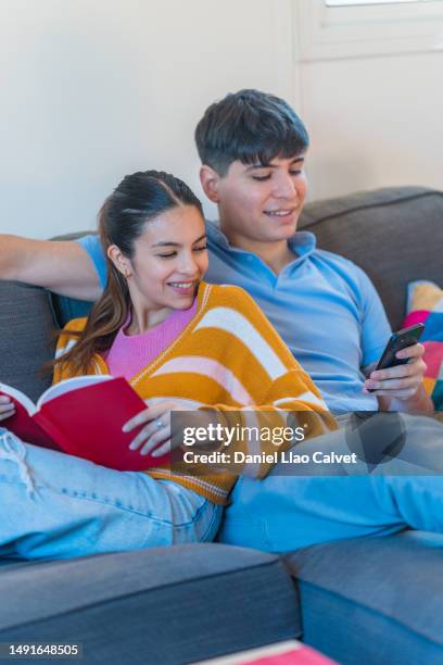 young couple with a mobile phone and a book while relaxing on the sofa at home. - casa calvet stock pictures, royalty-free photos & images