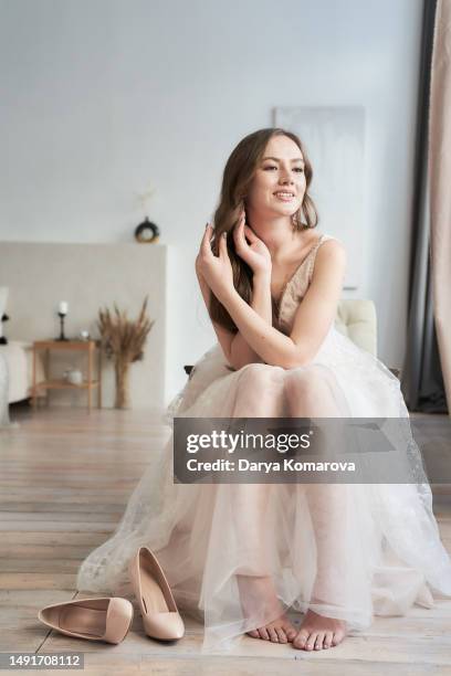beautiful young woman with long brown hair in wedding dress without shoes is sitting on the armchair on the background of cozy room with copy space. the happy woman is smiling. the concept of tired feet during the wedding day. - female foot models ストックフォトと画像