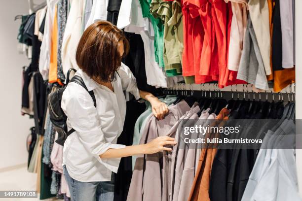 female customer in white blouse examining beige sweaters hanging on clothes rack - clothes hanging on rack at store for sale foto e immagini stock