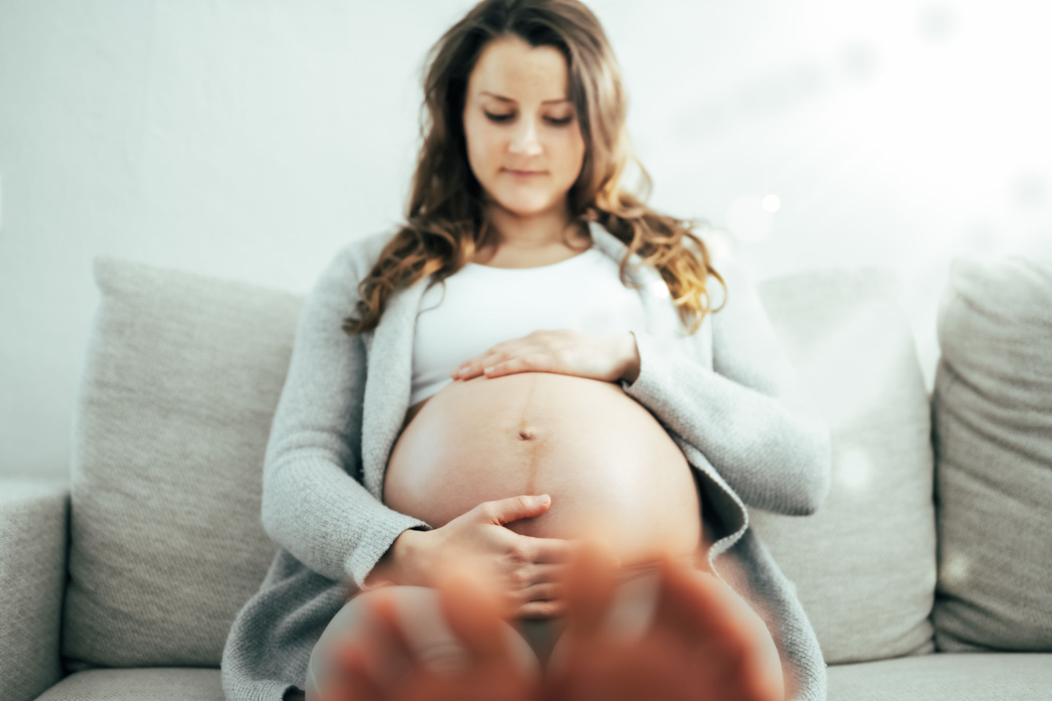 Frontal view of happy woman sitting on sofa gently holding her belly in imagination of baby in last stage of pregnancy. Pregnancy third trimester - week 34. Frontal view of happy woman sitting on sofa gently holding her belly in imagination of baby in last stage of pregnancy. Pregnancy third trimester - week 34.