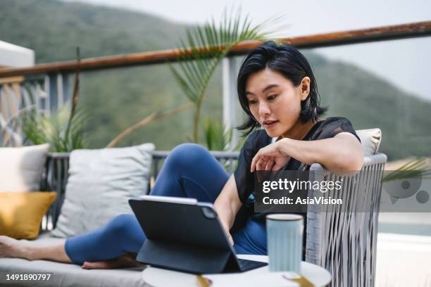 joven asiática sentada en el balcón, usando una tableta digital y tomando una taza de café por la mañana. mujer joven que usa la computadora para el ocio o los negocios en casa - informe de cuenta fotografías e imágenes de stock