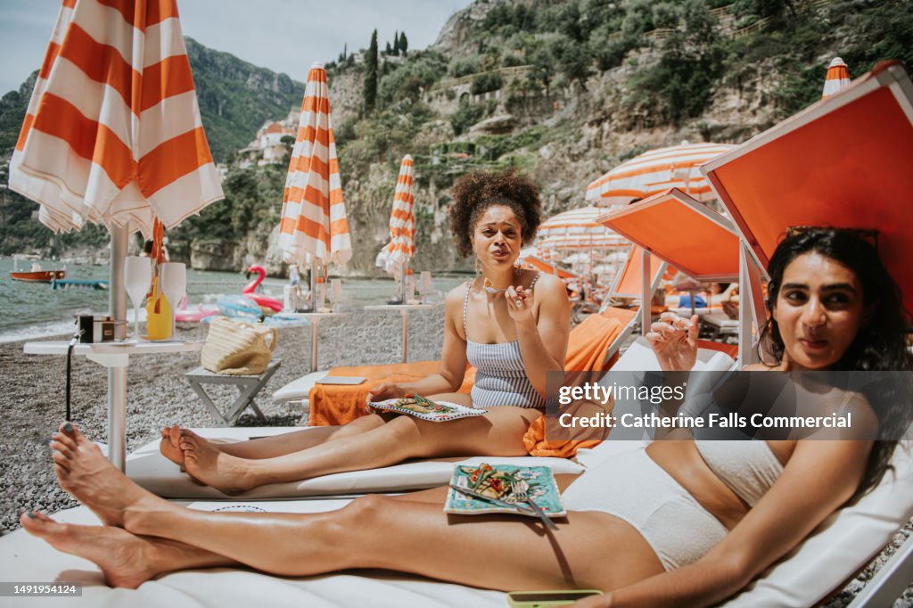 Two young woman enjoy a luxurious beach experience, eating lunch on a sun lounger.