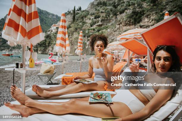 two young woman enjoy a luxurious beach experience, eating lunch on a sun lounger. - swim suit stock pictures, royalty-free photos & images