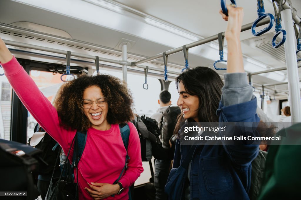 Two confident, beautiful woman stand on a bus, gripping the ceiling-mounted handles to steady themselves