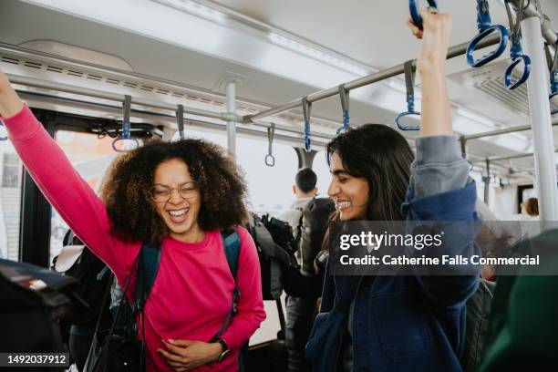 two confident, beautiful woman stand on a bus, gripping the ceiling-mounted handles to steady themselves - hauptverkehrszeit stock-fotos und bilder