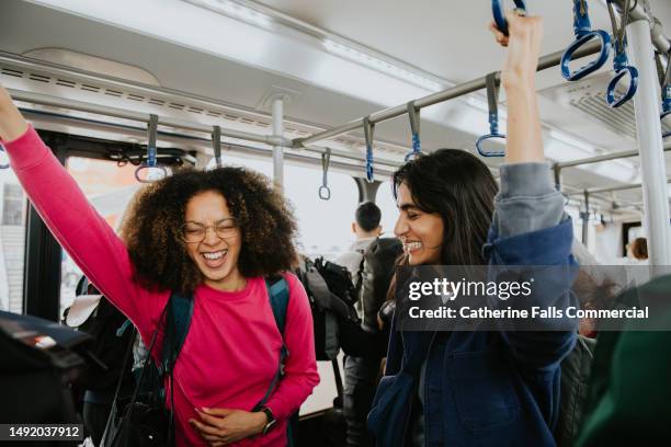 two confident, beautiful woman stand on a bus, gripping the ceiling-mounted handles to steady themselves - crowded bus stock pictures, royalty-free photos & images