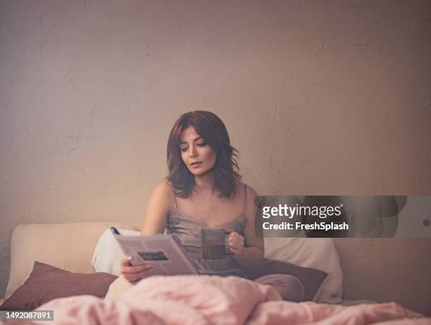 schöne rothaarige frau, die in nachtwäsche auf ihrem bett sitzt, eine zeitschrift liest und kaffee trinkt - splash news stock-fotos und bilder