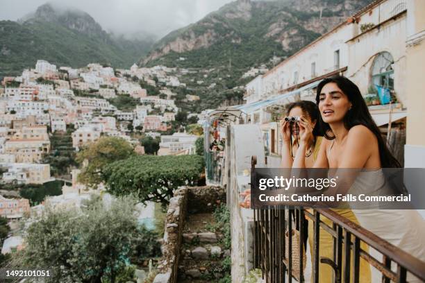 two beautiful woman perch on a railing and look out over the epic view of positano, italy. - indian tour guide stock pictures, royalty-free photos & images