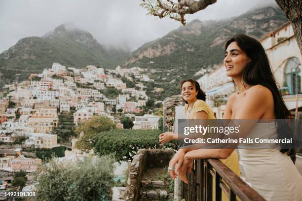 two beautiful woman perch on a railing and look out over the epic view of positano, italy. - local landmark stockfoto's en -beelden