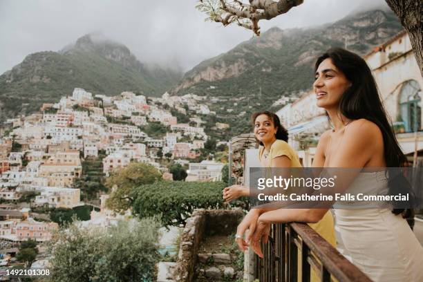 two beautiful woman perch on a railing and look out over the epic view of positano, italy. - ponto turístico local imagens e fotografias de stock