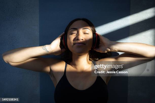 woman wearing headphones lying on yoga mat in sunbeams with arms outstretched and hands on ears - zen-like stock pictures, royalty-free photos & images