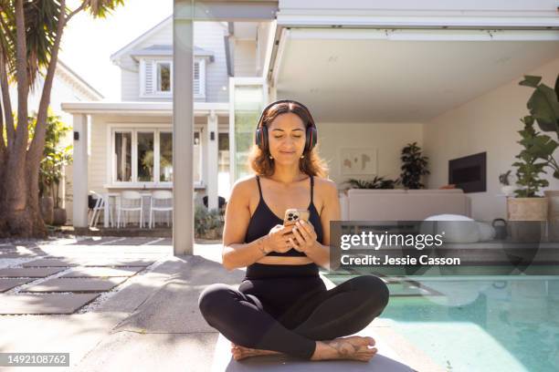 long haired woman wearing headphones and exercise wear sitting cross legged sitting beside swimming pool using mobile telephone - polynesische etniciteit stockfoto's en -beelden