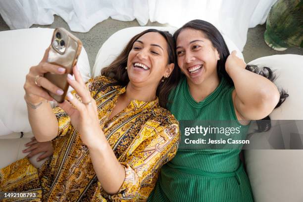 two long haired laughing women lying on sofa together using mobile telephone - polynesische etniciteit stockfoto's en -beelden