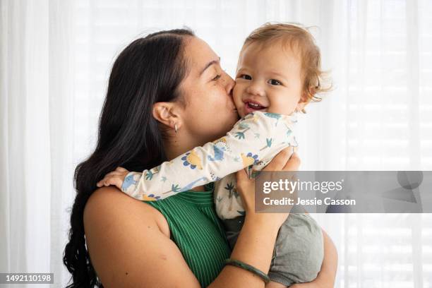 longhaired woman kissing smiling child's cheek in front of curtained window - maternity leave stockfoto's en -beelden
