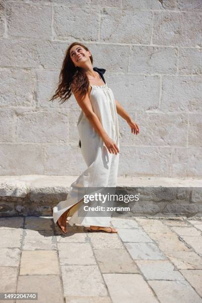 beautiful brunette wearing long sleeveless dress smiling over shoulder by stone brick wall in summer - sleeveless dress stock pictures, royalty-free photos & images