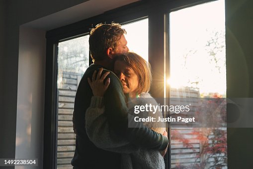 Powerful shot of a mature couple cuddling by the window in the late afternoon sun - emotional support Powerful shot of a mature couple cuddling by the window in the late afternoon sun - emotional support