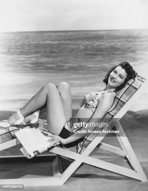 Ava Gardner poses on lounger wearing bikini on beach in a 1947 publicity portrait.
