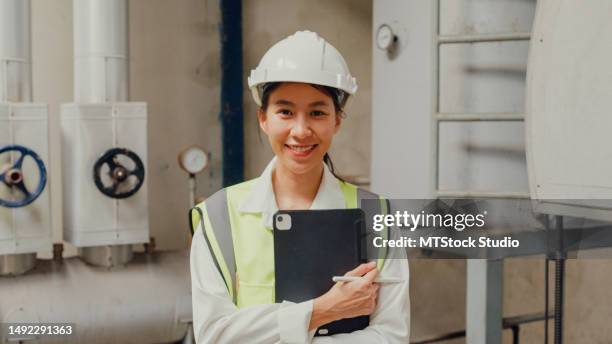 young asian female industrial engineer in hard hats smiling and looking at camera near steam boiler in chemical manufacturing factory. heavy industry and engineers. - camera operator imagens e fotografias de stock