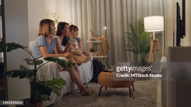 young asian women friends on sofa eat popcorn funny laugh watch comedy channel movie or sitcoms show on television in streaming online in home at night. leisure activity, lifestyle. - tv show stockfoto's en -beelden