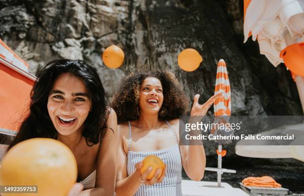 two beautiful woman juggle oranges on a sunny beach - italian beach fun stock pictures, royalty-free photos & images