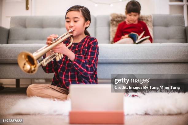 cheerful girl practising trumpet using the sheet music on digital table while her lovely sister reading book in background at home - wind instrument stock pictures, royalty-free photos & images