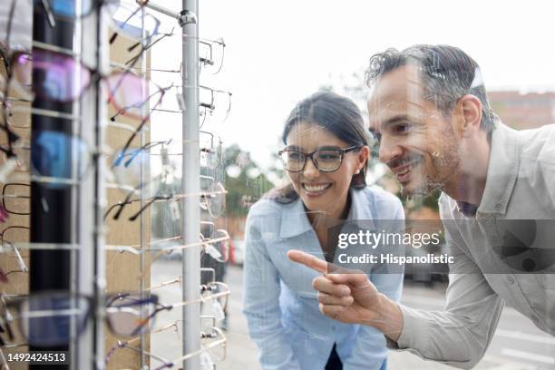 paar, das auf das schaufenster eines optikers schaut und auf gläser zeigt - schaufensterbummel stock-fotos und bilder