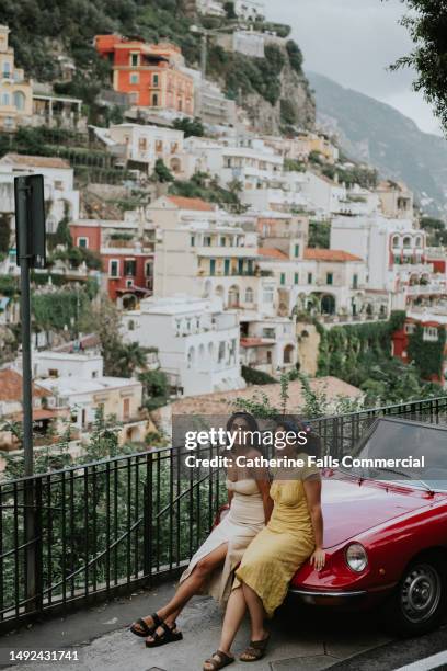 two beautiful young woman perch on the bonnet of a vintage red alfa romeo, with a view of positano in the background - indian tour guide stock pictures, royalty-free photos & images