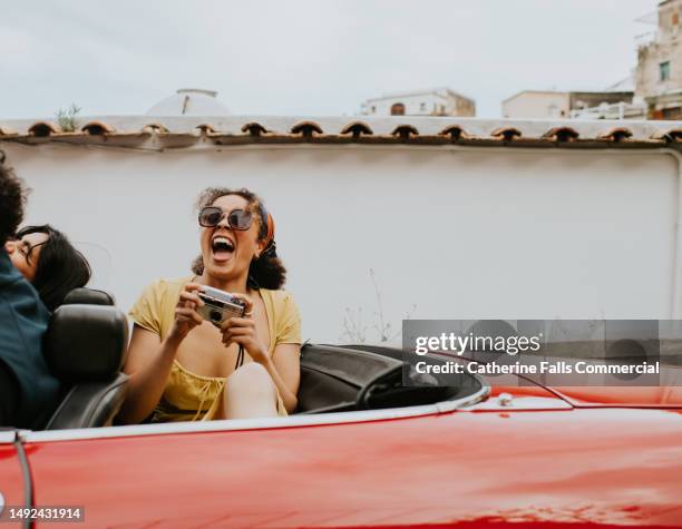 a pretty young woman sits in the back of a vintage red convertible and takes photos with a retro film camera - turismo esperienziale foto e immagini stock