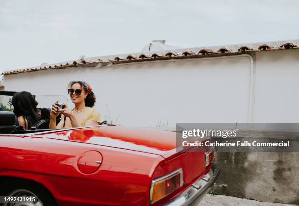 a pretty young woman sits in the back of a vintage red convertible and takes photos with a retro film camera - car photos stock-fotos und bilder