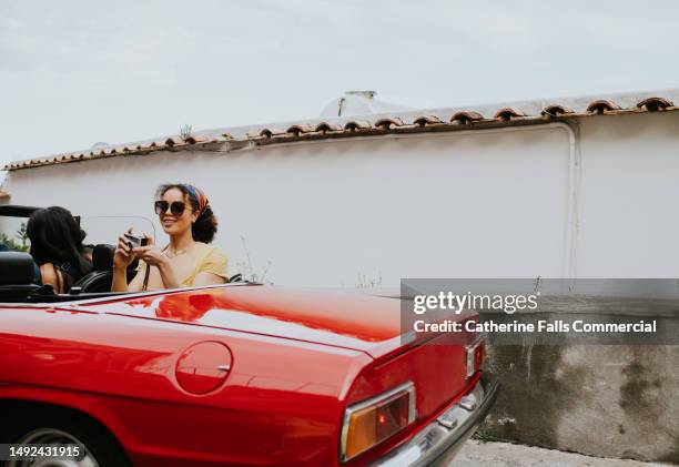 a pretty young woman sits in the back of a vintage red convertible and takes photos with a retro film camera - onboard kameraaufnahme stock-fotos und bilder
