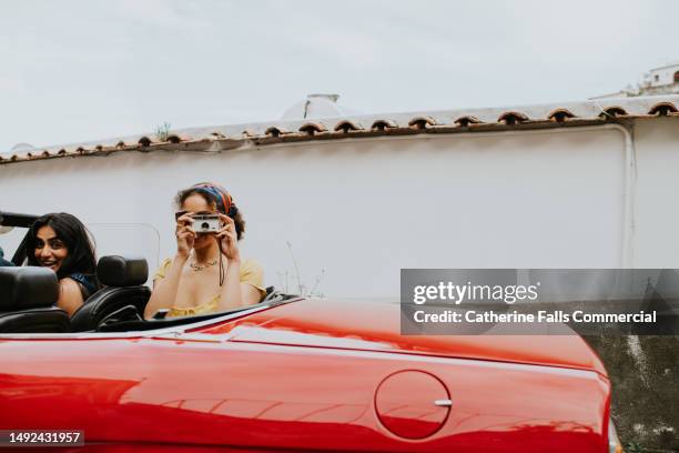 a pretty young woman sits in the back of a vintage red convertible and takes photos with a retro film camera - freedom to the camera stock pictures, royalty-free photos & images