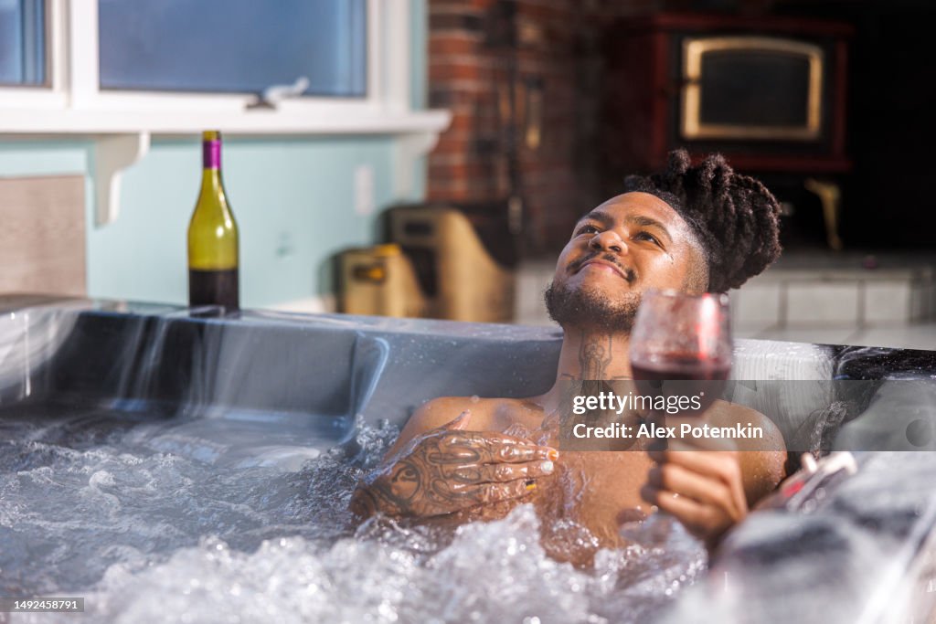 Freedom and relax in hot tub with wine. Joyful Dominican man with black dreadlocks and tattoo relax with wine glass in bubble bath