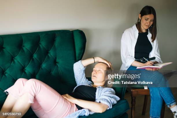 female therapist sitting on chair and writing while female patient lying on sofa with eyes closed relieving trauma - psychiatrists couch fotografías e imágenes de stock