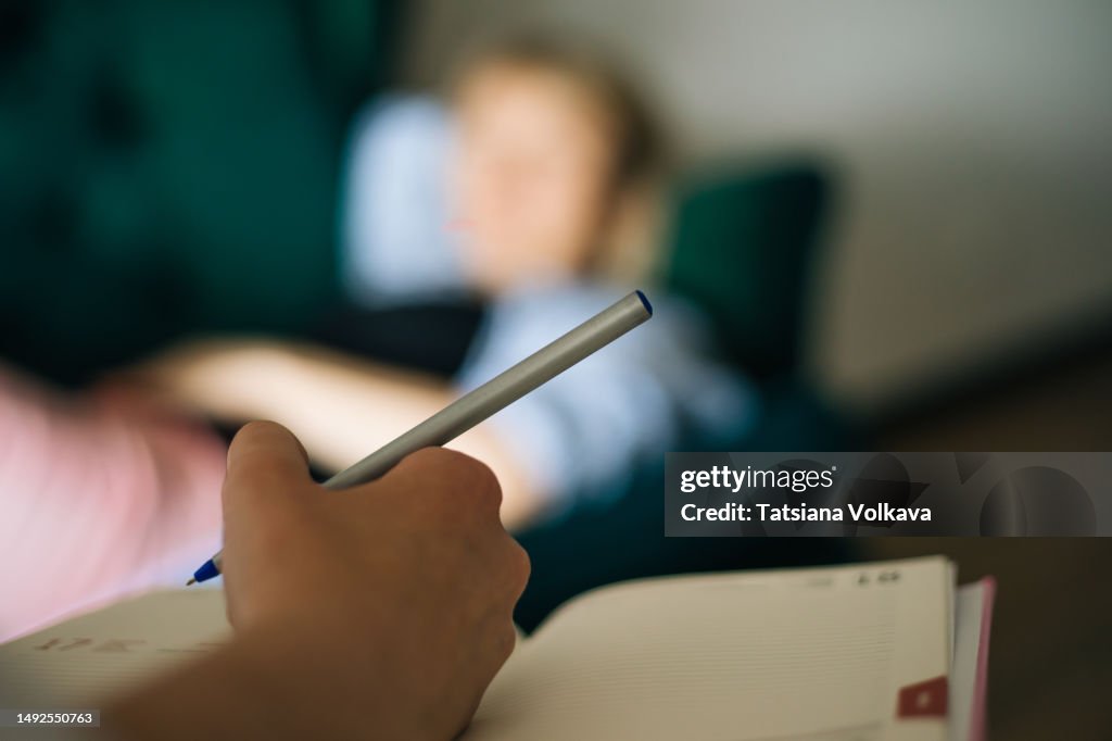 Hand of unrecognizable woman holding pen and notebook with blurred woman lying on sofa in background