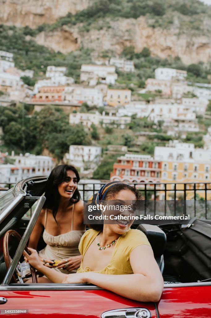 Two woman inside a vintage red convertible with a view of Positano, Italy behind them