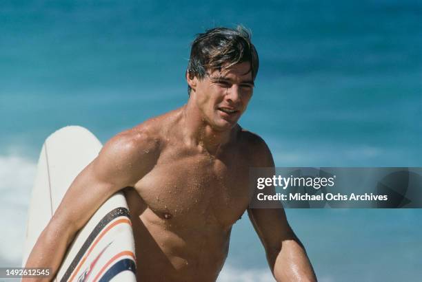 American actor Jan-Michael Vincent a surfboard under his right arm on the set of 'Big Wednesday', at Sunset Beach, on the north shore of Oahu ,...