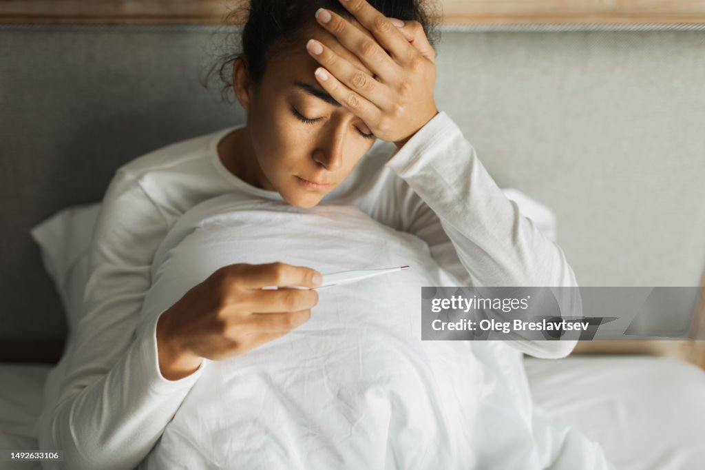 Woman with headache and fever taking her temperature with thermometer. Bad health condition