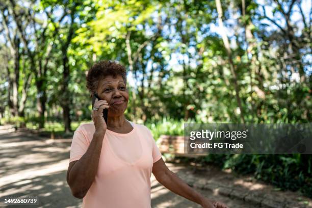 senior woman talking on the mobile phone while walking in the park - senior women walking stock pictures, royalty-free photos & images