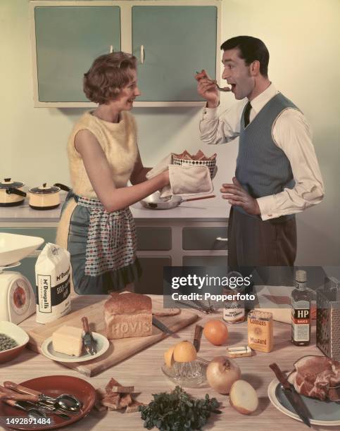 Posed studio portrait of a woman and man preparing and cooking food in a kitchen setting, the man uses a spoon to taste a dish prepared by the woman...